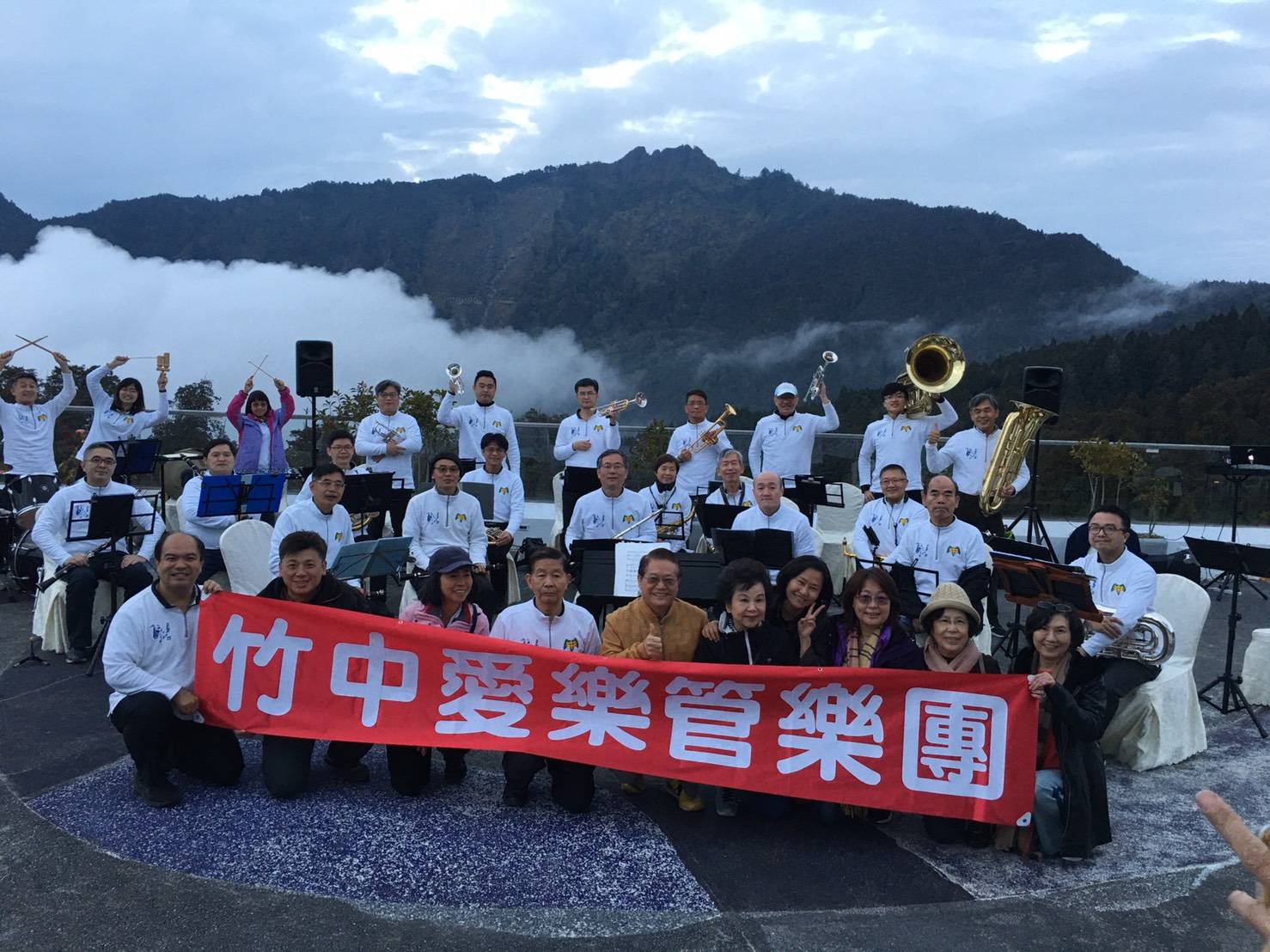 竹中愛樂管樂團創團首次遠征阿里山演出的全團大合照，團員穿著白色團服、拉著紅色團名布條，背景是雲霧繚繞的山景